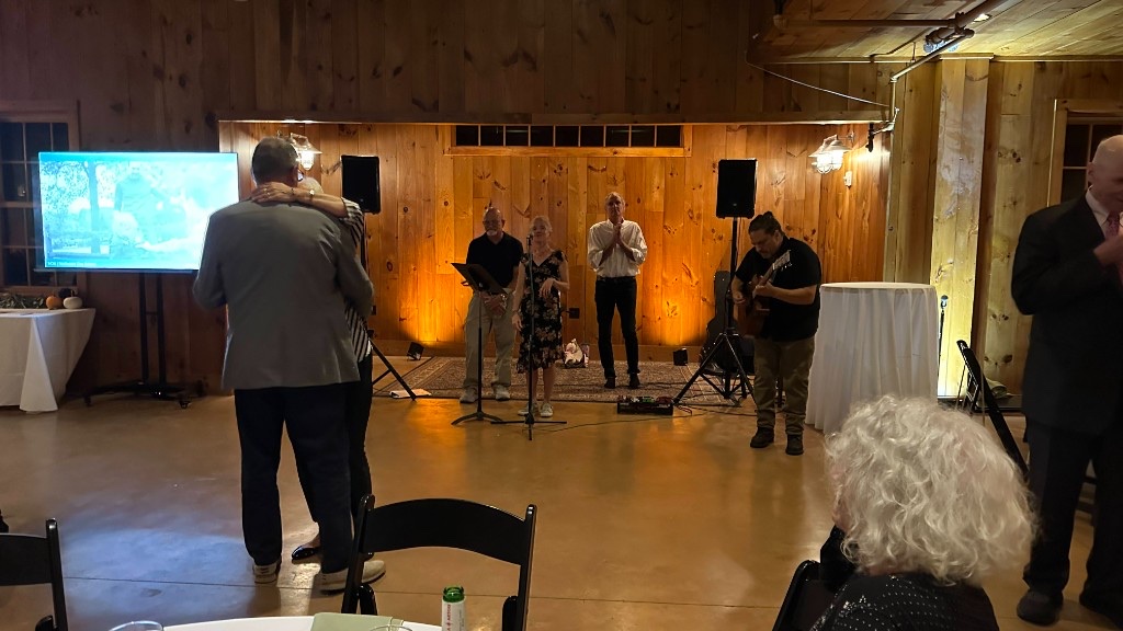 Guests at the microphones in front of a 75-inch, with warm uplight on the barn wall.