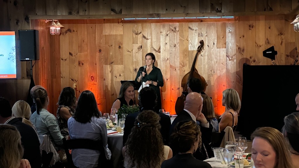 A speaker at a wireless microphone during the Harvest Moon Gala, with uplighting on the barn wall.