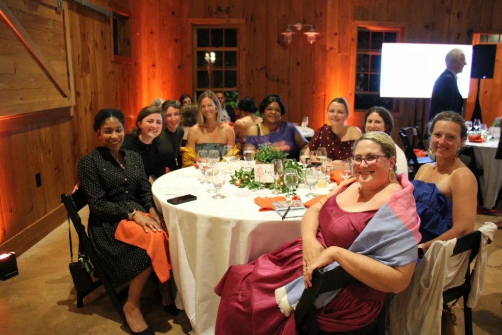 Guests at a round table in the Celebration Barn, with amber uplighting and a 75-inch in the background.